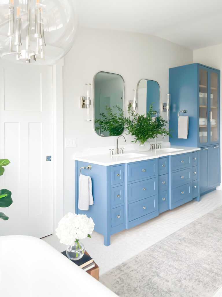 A primary bathroom design featuring classic and contemporary elements. The custom blue vanity including a linen closet is balanced by white quartz countertops, light gray walls, and white tile. #SherwinWilliamsReservedWhite #BenjaminMooreLazySunday #BlueBathroomVanity #PaintColor #LightGrayBathroomWallsWithBlueVanity #dbCPHistoricEdgefieldProject